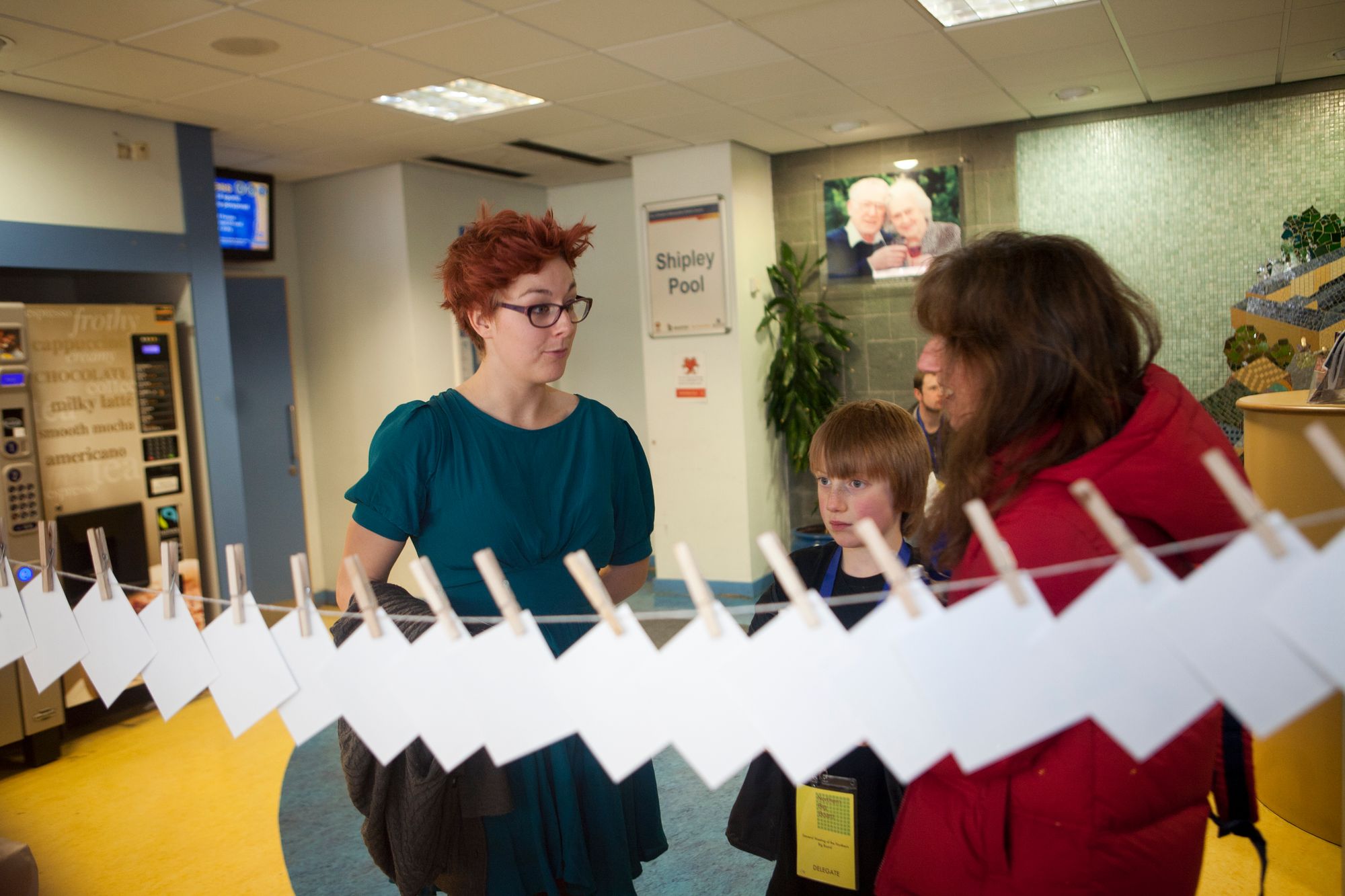 A young woman with red spiky hair and a blue dress on talks to a woman in a red parka in the lobby of a swimming pool, in the front of the shot are a number of cards pegged to a piece of string, they are photographed from the blank side.