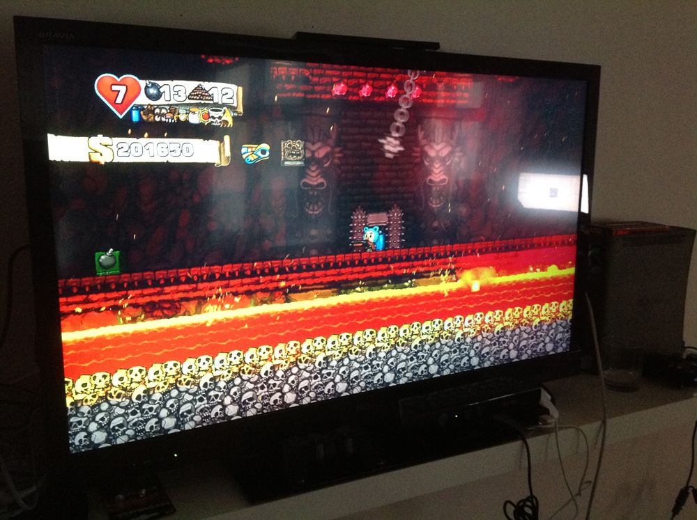 A photo of a tv screen showing the videogame Spelunky. A player character with a shotgun enters Yama's Lair. There's a big bomb box on the left.