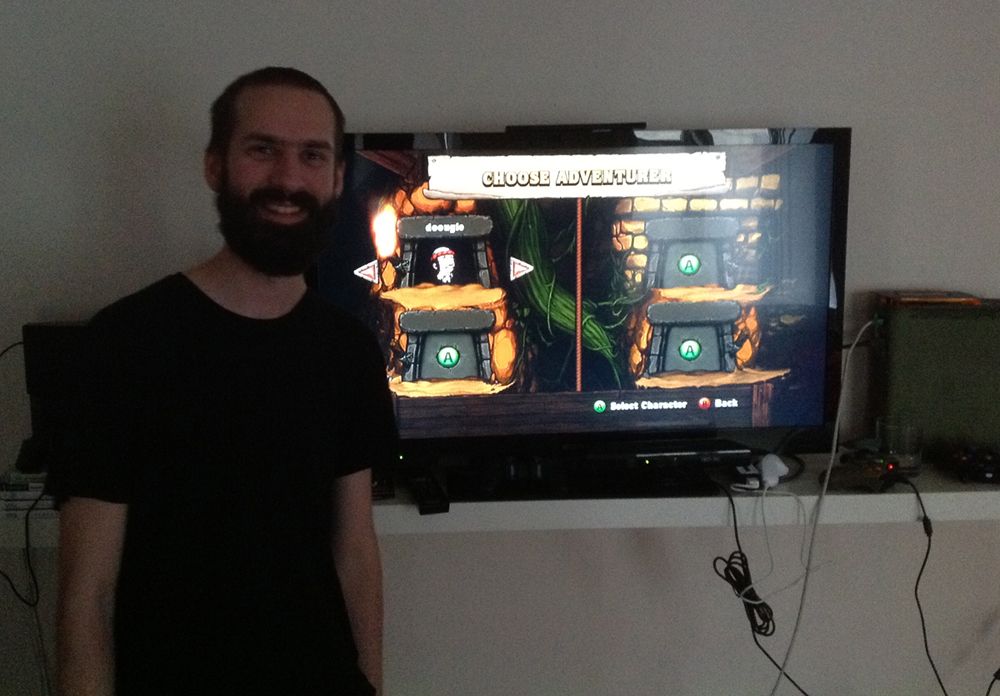 A photo of Swedish developer Nicklas "Nifflas" Nygren standing in front of a television showing the videogame Spelunky. The screen is on the CHOOSE ADVENTURER screen, with the avatar Yang currently selected.