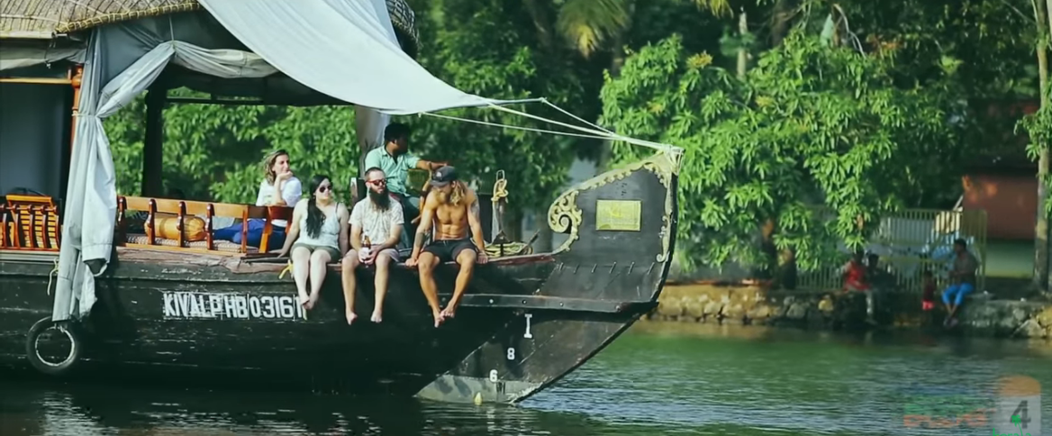 Three people dangle their legs over the edge of a boat on a river. Another person is leaning across railing and another person drives the boat.