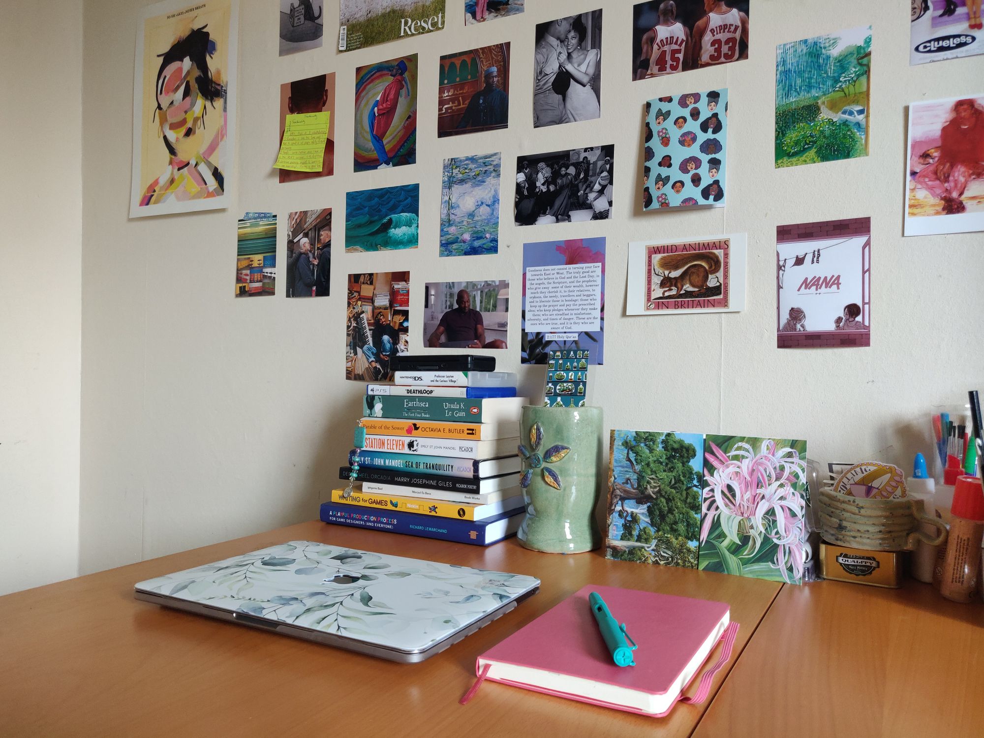 Halima's work desk with laptop, notebook and pen. Stacks of books are on the desk as well as stationery and colourful posters are on the wall.