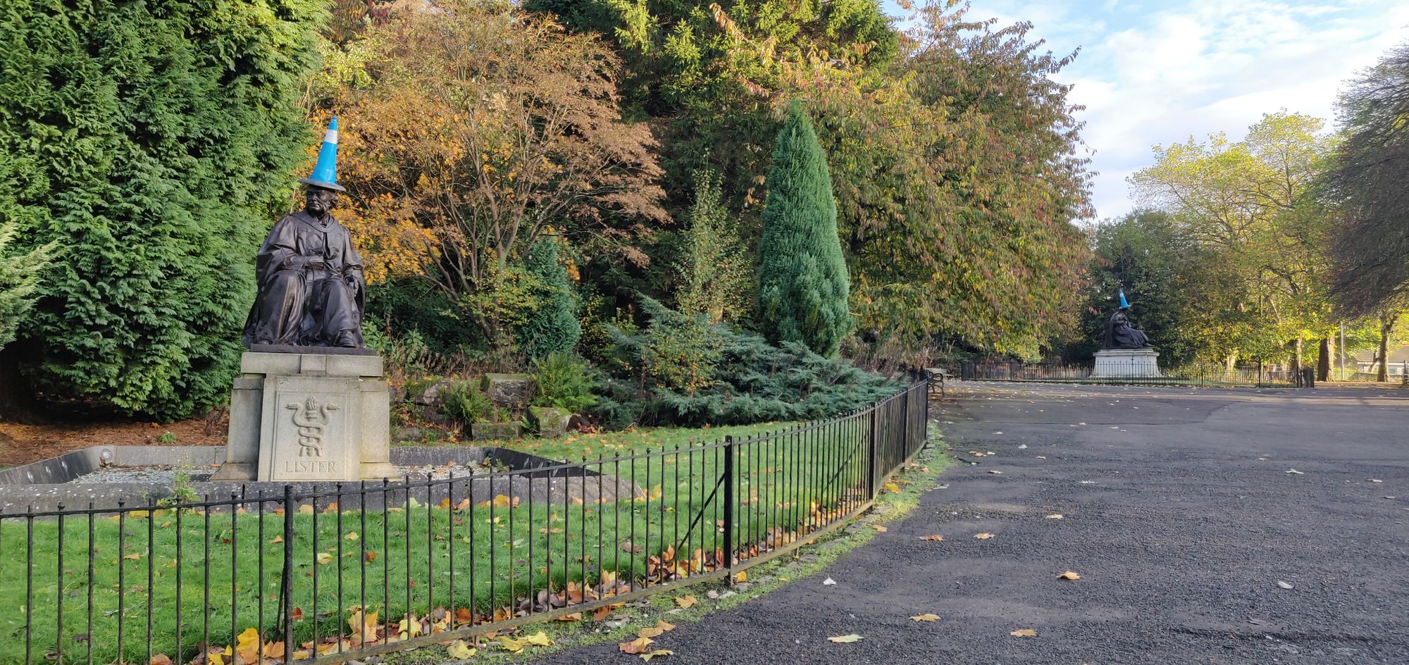 An image taken in a park. On the left is seated statue of a person with a blue and white cone place on top of it. There are green and orange leaved trees in the background. In the distance on the right is another statue similar to the left one with also a cone on its head.