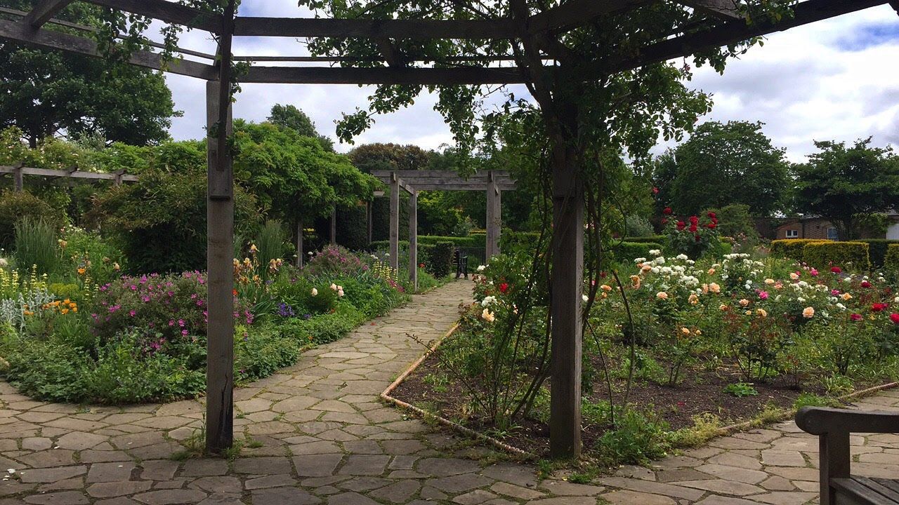 An image from the Botanical space in Brockwell Park, Brixton, London. The image has two beds filled with pink, orange, white, red and purple flowers. There is a stone pathway that divides the flowerbeds and there are also two wooden pergolas.