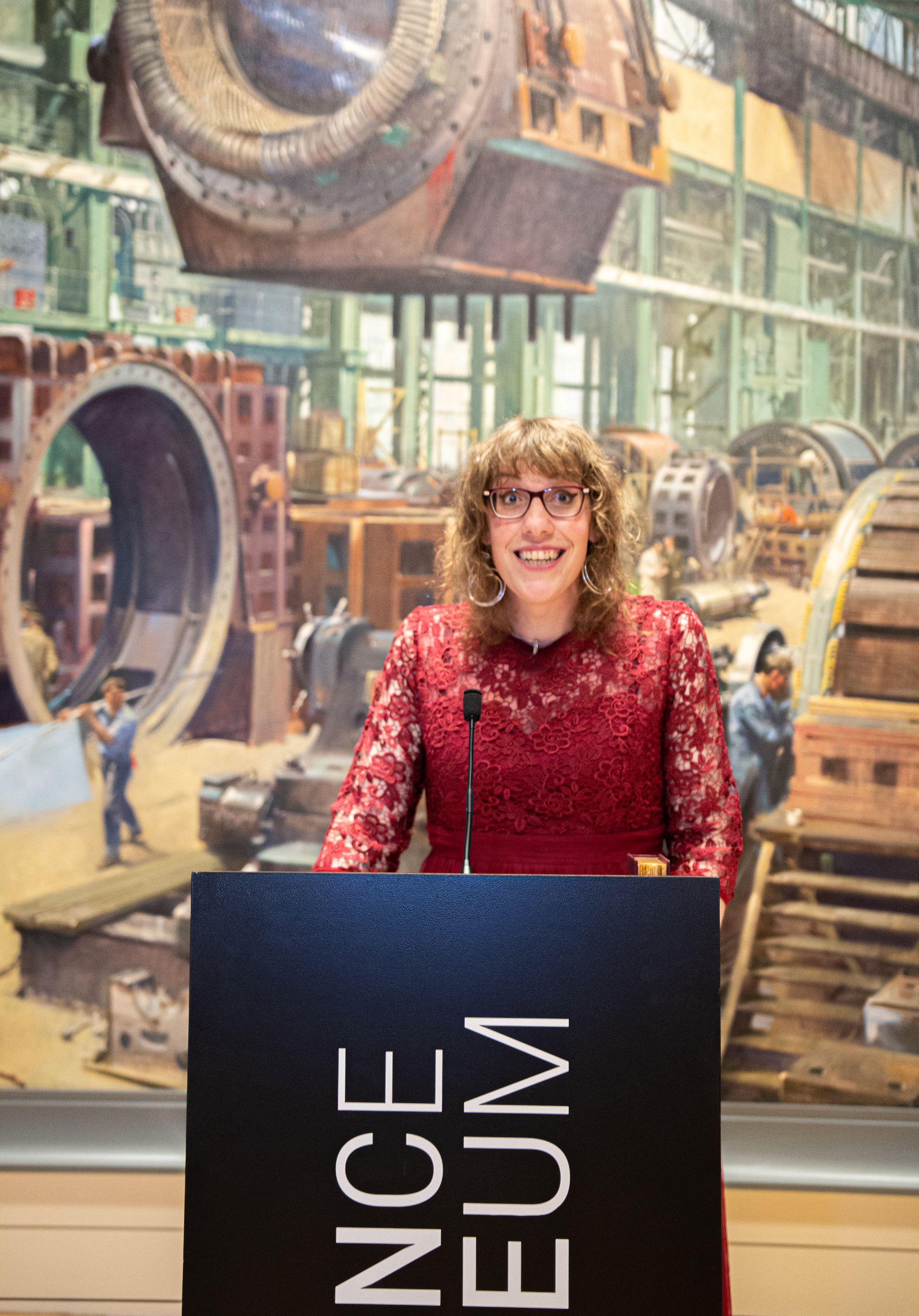 Harry Josephine Giles wearing a red dress, stands at a black podium at the Science Museum accepting her Arthur C. Clarke Award. The background is a colourful mural featuring people working on various machinery.