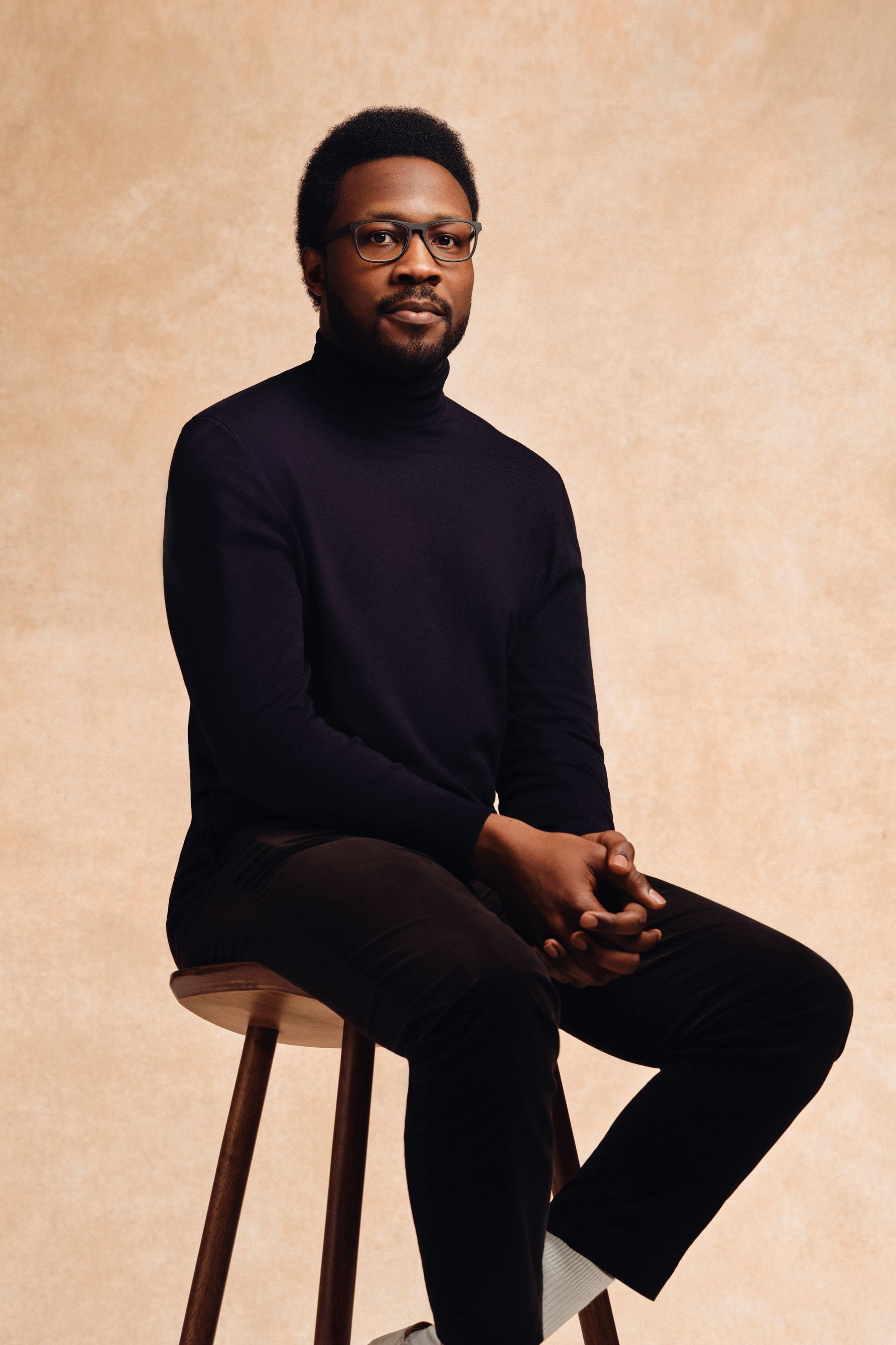 Ben Wilson sits on a stool with a pale background. He is wearing a black turtleneck and trousers.