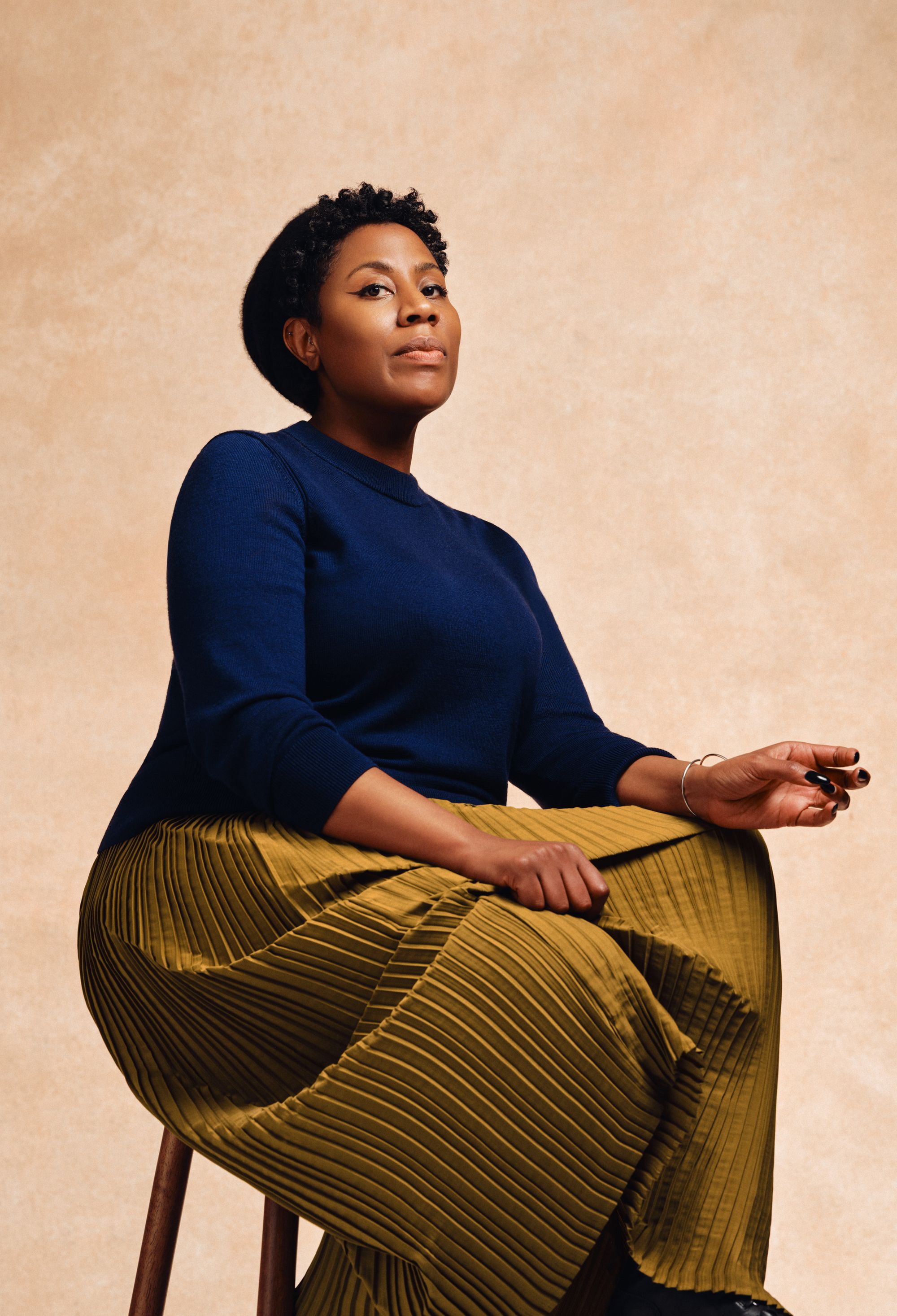Sharna Jackson sits on a stool with a pale background. She wears a dark blue jumper and a golden long skirt that goes to her ankles.